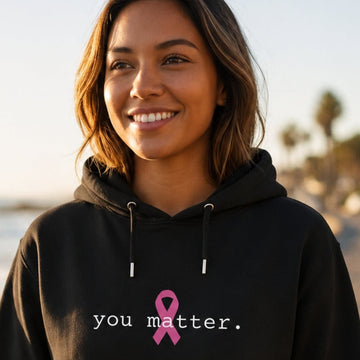 A woman wearing a black hoodie with the text 'you matter.' and a pink awareness ribbon printed on the chest, standing on a coastal road with the ocean in the background.