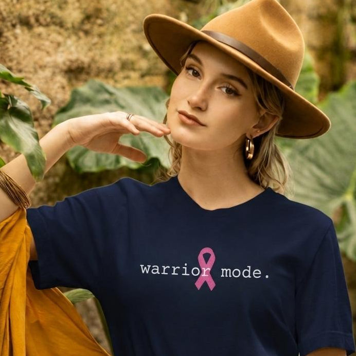 A woman wearing a navy blue graphic t-shirt with the text 'warrior mode.' and a pink ribbon, styled with a wide-brimmed tan hat, a yellow-orange draped jacket, and a white skirt with a large brown belt, set against a lush green tropical background.