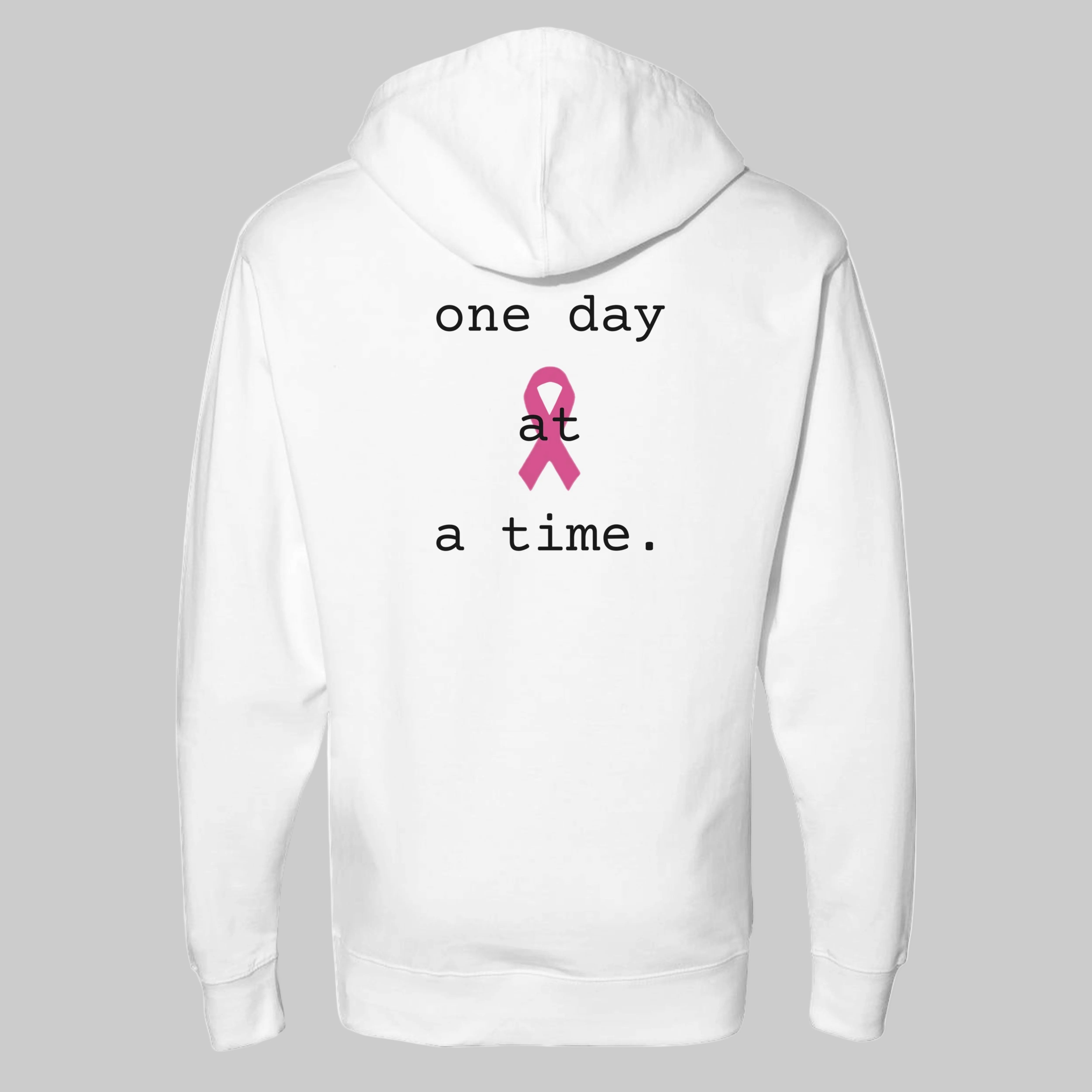 White pullover hoodie shown from the back, featuring the printed text 'one day at a time.' in black around a pink awareness ribbon graphic on a warm gray-colored background.
