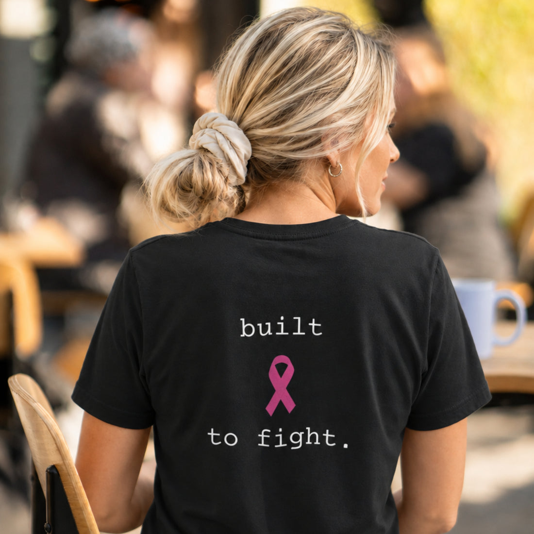 A woman wearing a black short-sleeve t-shirt with the text 'built to fight.' in white and a pink awareness ribbon graphic, photographed in a cafe setting.