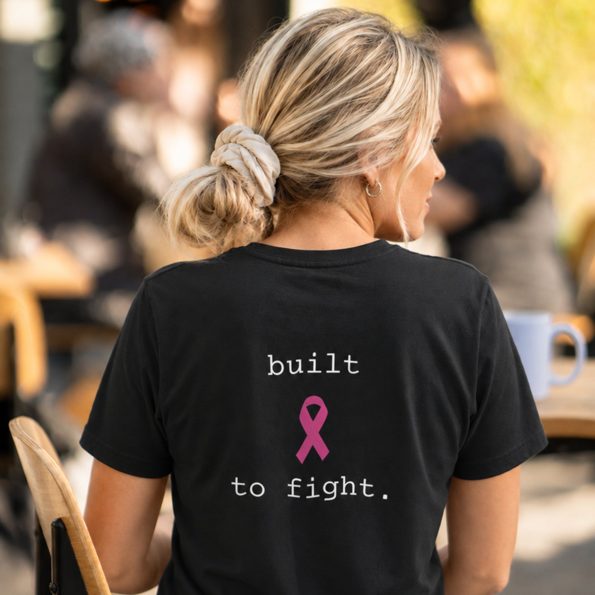 A woman viewed from behind wearing a black short-sleeve T-shirt with the words 'built to fight.' in white and a pink ribbon graphic on the back, photographed in a cafe setting.